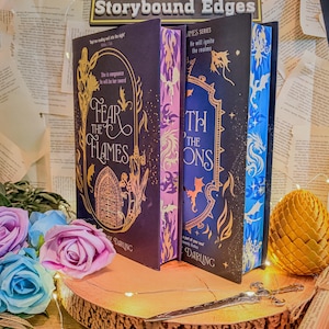 May include: A display of fantasy books with decorative covers. The books are arranged on a wooden slice with a sword and flowers. The book spines are visible, with titles like "Fear the Flames" and "Thorns". The background features pages and fairy lights.