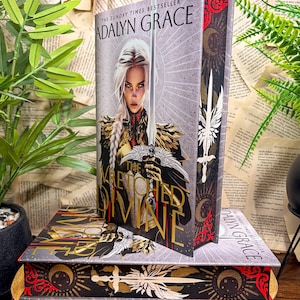 May include: A hardcover book titled The Wretched Divine by Adalyn Grace. The cover features a blonde character holding a sword, with gold and red celestial motifs. The spine and back cover display a white sword, red filigree, and moon symbols.