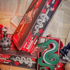 Op de afbeelding: Een stapel boeken met de titel "Terror of the Gates" en een decoratieve groene slangenhanger. De boekomslagen zijn zwart met rode en witte accenten. Een kleine, sierlijke parfumfles is ook zichtbaar.