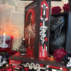 Op de afbeelding: Een boek getiteld "Terror at the Gates" van Scarlett St. Clair met decoratieve randen met een zwaard- en slangontwerp. De afbeelding bevat een zwart drakenbeeld, een parfumfles en een kaars. De tekst "Storybound Edges" is zichtbaar.