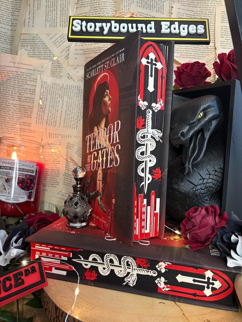 Op de afbeelding: Een boek getiteld "Terror at the Gates" van Scarlett St. Clair met decoratieve randen met een zwaard- en slangontwerp. De afbeelding bevat een zwart drakenbeeld, een parfumfles en een kaars. De tekst "Storybound Edges" is zichtbaar.