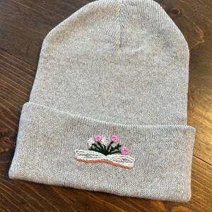 May include: Gray knit beanie with an embroidered design of a book with pink flowers growing out of it.