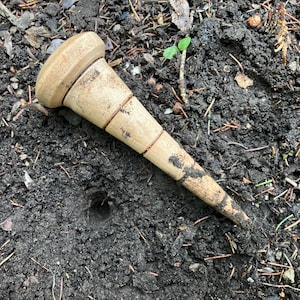 May include: A light brown wooden tool with a rounded top and a tapered, pointed end. The tool has several dark brown bands and is partially covered in soil. It is lying on dark soil.