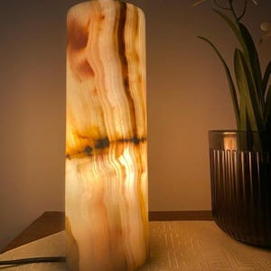 May include: A cylindrical onyx lamp with a warm, glowing interior. The lamp's exterior features a natural pattern of beige, brown, and white striations. The lamp is illuminated, casting a soft light. A dark vase with greenery is to the right.