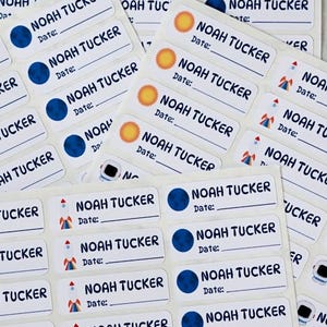 May include: Sheets of personalized name labels with the name "NOAH TUCKER" printed on each. The labels feature space-themed graphics like planets, rockets, and astronauts, with a space for the date.