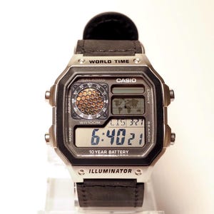 Puede incluir: Un reloj digital Casio con una correa de cuero negro. La esfera del reloj muestra la hora, la fecha y las funciones de hora mundial. El reloj tiene una caja plateada y el texto "CASIO", "WORLD TIME" e "ILLUMINATOR" son visibles.