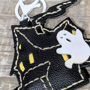 May include: Black leather Halloween-themed keychain featuring a house shape with yellow window accents and a white ghost. The keychain has a white clasp and is stitched with cream-colored thread.