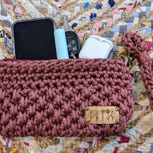May include: A crocheted burgundy zippered pouch with a brown leather tag that says "Pretty." The pouch has a long burgundy crocheted strap. The pouch is open and contains a black phone, a blue tube, a key, and a white case.