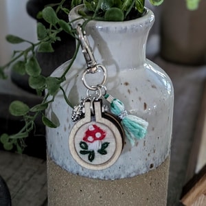 May include: A handmade keychain featuring a miniature embroidered mushroom design. The keychain includes a wooden hoop, a silver-tone clasp, and a small tassel with a light blue and silver color scheme. The background is a ceramic vase with greenery.