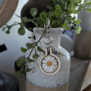 May include: A wooden embroidery hoop keychain featuring a white daisy with a yellow center. The keychain has a silver clasp and small charms. The background includes a ceramic vase with greenery.