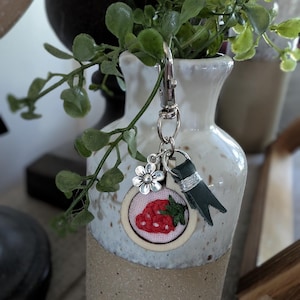 May include: A handmade keychain featuring a small, embroidered strawberry design within a wooden hoop. The keychain includes a silver-tone clasp, a flower charm, and a dark green leather accent. The background shows a ceramic vase with greenery.