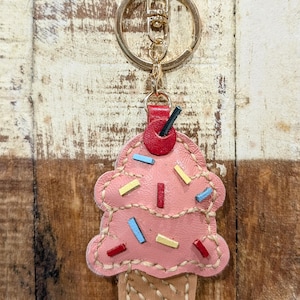 May include: Pink leather cupcake keychain with a gold-tone keyring. The cupcake has a stitched design with colorful rectangular sprinkles and a red cherry. The cone is tan and stitched.