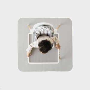 May include: A white high chair with a gray and white patterned seat cushion. A child is sitting in the chair. The chair is on a gray mat.