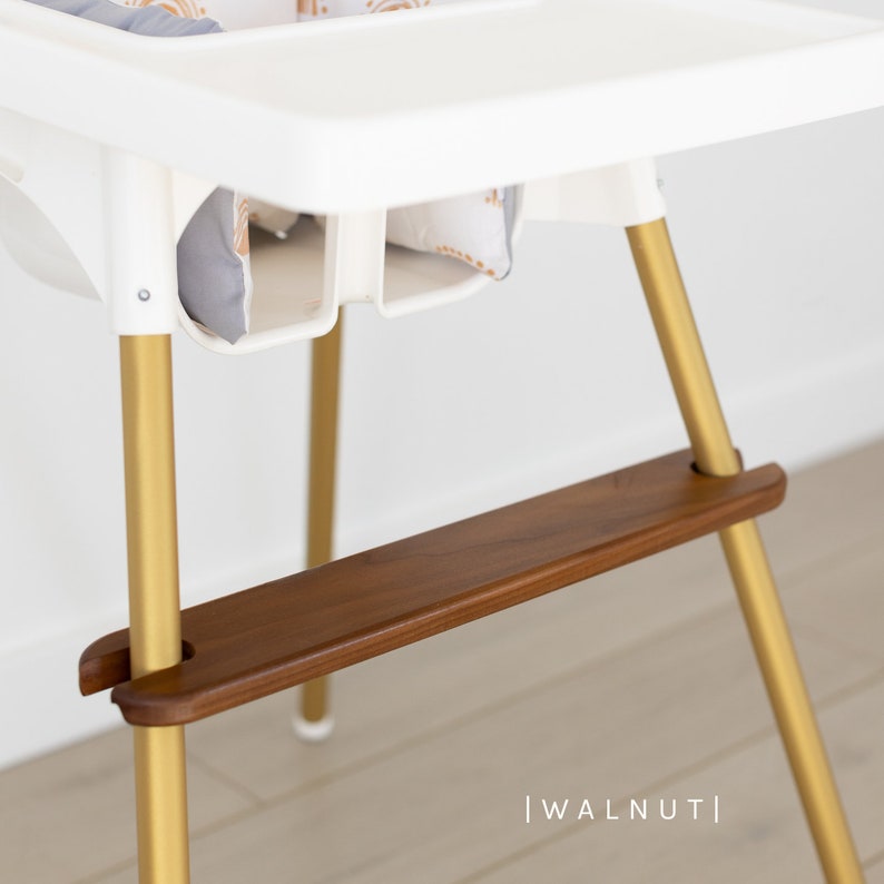 May include: A white high chair with gold-colored legs and a walnut-colored footrest. The chair has a white tray and a patterned cushion. The text "| WALNUT |" is at the bottom of the image.