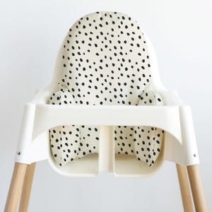 May include: A white high chair with a cream-colored seat and backrest featuring a black polka dot pattern. The chair has wooden legs and a tray for a baby or toddler.