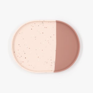 Puede incluir: Una bandeja redonda de color rosa y marrón con textura moteada. La bandeja está dividida en dos, con un lado de color marrón sólido y el otro lado con una textura rosa moteada.