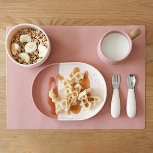 May include: A pink placemat holds a breakfast setting. A bowl of cereal with banana slices, a mug of milk, a plate of waffles with syrup, and a fork and spoon are arranged on the mat. The plate is white with a speckled design.