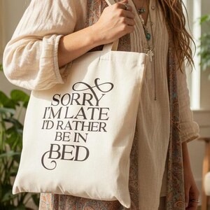May include: A cream canvas tote bag with the text "Sorry I'm late I'd rather be in bed" in black script. The bag is held by a person wearing a cream blouse and a patterned skirt. The background includes plants and a macrame wall hanging.
