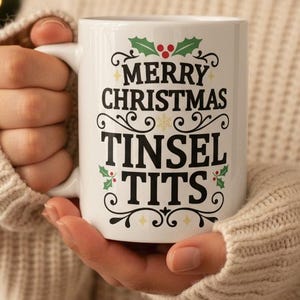 May include: White ceramic mug with the text "MERRY CHRISTMAS TINSEL TITS" in black and green. The mug features holly and berry accents. The mug is held by a person wearing a cream-coloured sweater.