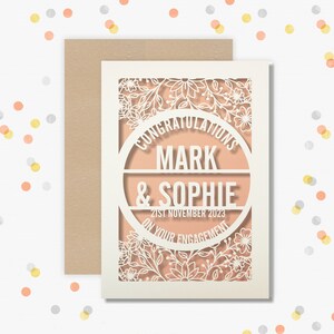 May include: A white card with a delicate floral design cut out. The card has a peach background with a circle in the center that says "Congratulations Mark & Sophie on your engagement 21st November 2023".