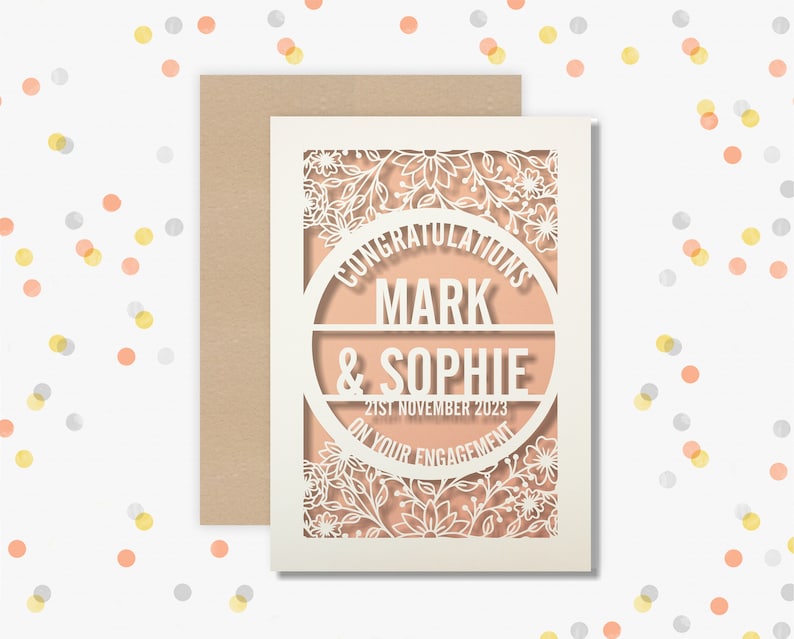 May include: A white card with a delicate floral design cut out. The card has a peach background with a circle in the center that says "Congratulations Mark & Sophie on your engagement 21st November 2023".