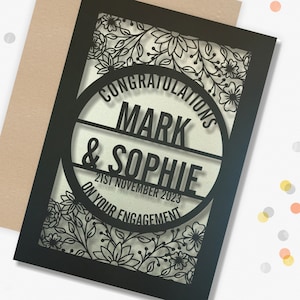 May include: Black laser cut card with a floral design. The card says "Congratulations Mark & Sophie on your engagement 21st November 2023".