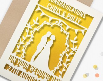 May include: A cream-colored wedding card with a laser-cut design. The card features the words "Congratulations Chris & Polly" and "On Your Wedding Day 27th August 2022". A silhouette of a couple is in the center, surrounded by floral and banner details.