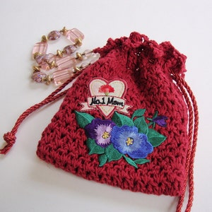 May include: Red crocheted drawstring bag with a floral embroidery design. The embroidery features two purple and blue flowers with green leaves. The bag also has a heart-shaped patch with the text "No. 1 Mom" in black.