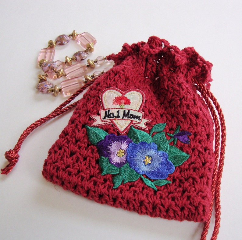 May include: Red crocheted drawstring bag with a floral embroidery design. The embroidery features two purple and blue flowers with green leaves. The bag also has a heart-shaped patch with the text "No. 1 Mom" in black.
