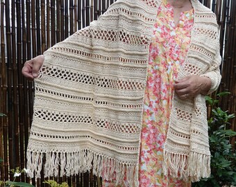 May include: A crocheted shawl with a delicate openwork pattern and fringe trim. The shawl is made in a cream color and is draped over a person wearing a floral dress.