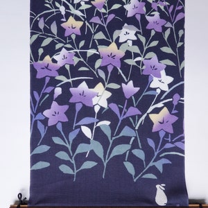 May include: A dark blue fabric wall hanging featuring a white rabbit and a floral pattern of purple, white, and yellow flowers. The fabric is rolled up on a wooden dowel with black cords.