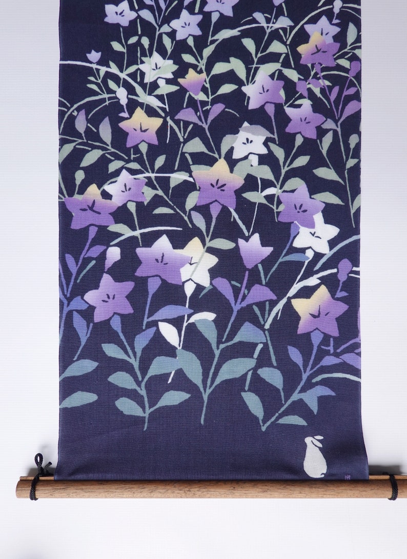 May include: A dark blue fabric wall hanging featuring a white rabbit and a floral pattern of purple, white, and yellow flowers. The fabric is rolled up on a wooden dowel with black cords.