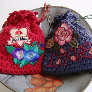 May include: Two crocheted drawstring bags with embroidered designs. One bag is red with a heart and the text "No. 1 Mom". The other bag is purple with a rose and the word "LOVE" embroidered in red.