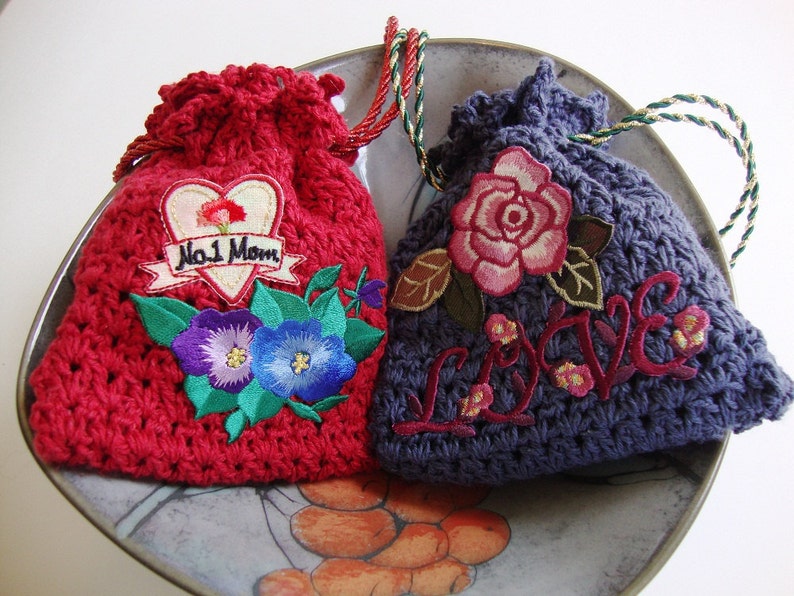 May include: Two crocheted drawstring bags with embroidered designs. One bag is red with a heart and the text "No. 1 Mom". The other bag is purple with a rose and the word "LOVE" embroidered in red.