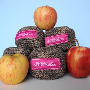 Op de afbeelding: Een stapel van vier bruine gehaakte ballen met roze labels waarop "Basket of Deplorables" staat. Drie rode appels liggen bovenop de ballen.