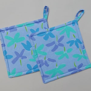 Puede incluir: Dos agarraderas de olla azules con un patrón floral en tonos de azul, verde y morado. Las agarraderas tienen un lazo para colgar.