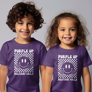 Op de afbeelding: Twee kinderen dragen paarse T-shirts met een witte geruite achtergrond en een glimlachend gezicht met vredestekens. De tekst op de shirts luidt "Purple Up Military Child".