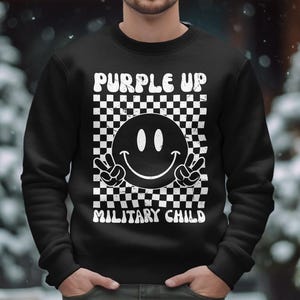 Op de afbeelding: Zwart sweatshirt met een wit geruit patroon, een smiley met vredestekens en de tekst "Purple Up Military Child".
