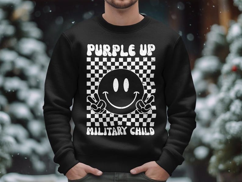 Op de afbeelding: Zwart sweatshirt met een wit geruit patroon, een smiley met vredestekens en de tekst "Purple Up Military Child".