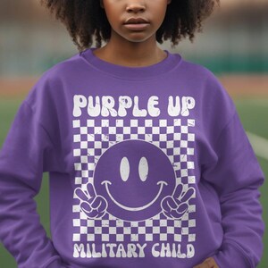 Op de afbeelding: Paarse trui met een wit geruit patroon, een smiley en de tekst "Purple Up Military Child".