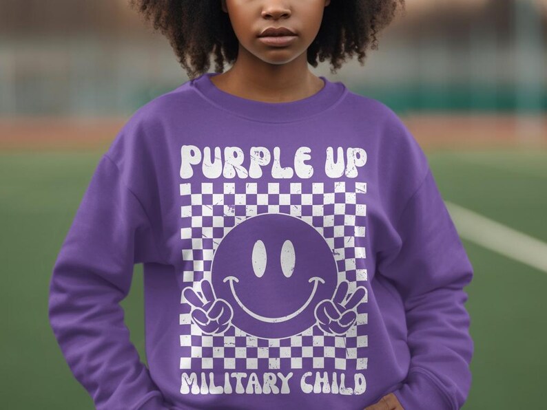 Op de afbeelding: Paarse trui met een wit geruit patroon, een smiley en de tekst "Purple Up Military Child".