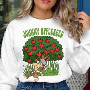 May include: White sweatshirt with a cartoon illustration of Johnny Appleseed planting an apple tree. The text "Johnny Appleseed" is printed in green letters on the front of the sweatshirt.