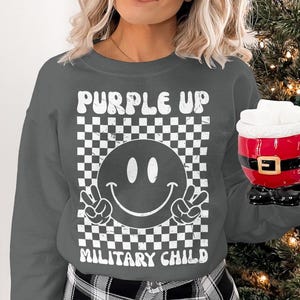 Op de afbeelding: Een grijs sweatshirt met een wit grafisch ontwerp. De grafiek bevat een geruit patroon, een smiley met vredestekens en de tekst "Purple Up Military Child".