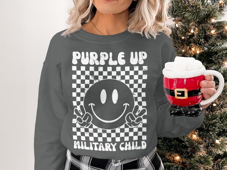 Op de afbeelding: Een grijs sweatshirt met een wit grafisch ontwerp. De grafiek bevat een geruit patroon, een smiley met vredestekens en de tekst "Purple Up Military Child".