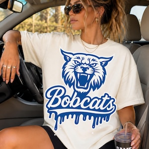 Op de afbeelding: Een crèmekleurig T-shirt met korte mouwen voorzien van een grote blauwe afbeelding van een grommende bobcatkop boven het woord "Bobcats" in een scriptlettertype, met een druipend verfeffect onderaan het tekstontwerp op de voorzijde.