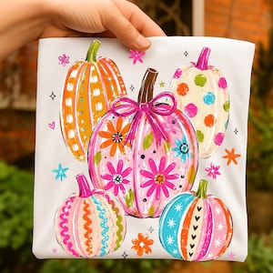 May include: A white t-shirt featuring a graphic print of five pumpkins in various colors and patterns. The central pumpkin is pink with floral designs and a pink bow. Other pumpkins feature orange stripes, colorful polka dots, and wavy lines.