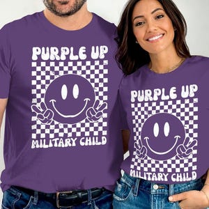 Op de afbeelding: Twee paarse T-shirts met een witte geruite achtergrond, een witte smiley met peace-tekens en de tekst "Purple Up Military Child".