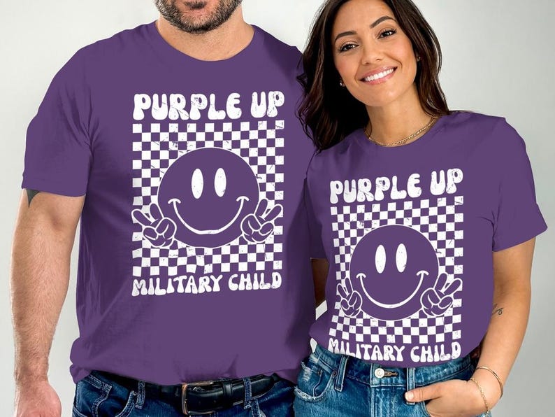 Op de afbeelding: Twee paarse T-shirts met een witte geruite achtergrond, een witte smiley met peace-tekens en de tekst "Purple Up Military Child".