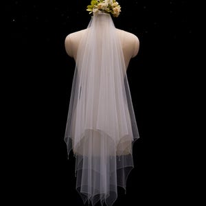 May include: A two-tier white tulle wedding veil featuring a scalloped edge with delicate bead embellishments along the hem. The veil is attached to a floral headpiece composed of small cream and pale yellow fabric flowers at the crown.