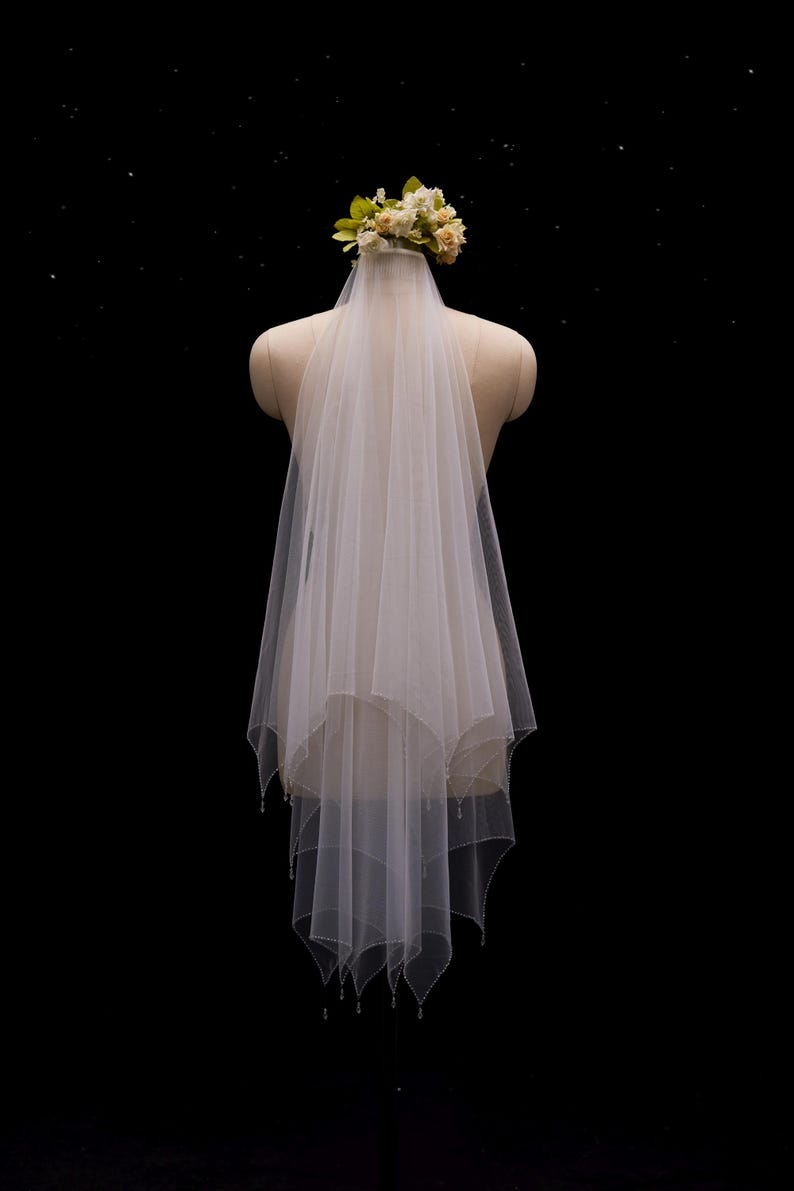 May include: A two-tier white tulle wedding veil featuring a scalloped edge with delicate bead embellishments along the hem. The veil is attached to a floral headpiece composed of small cream and pale yellow fabric flowers at the crown.